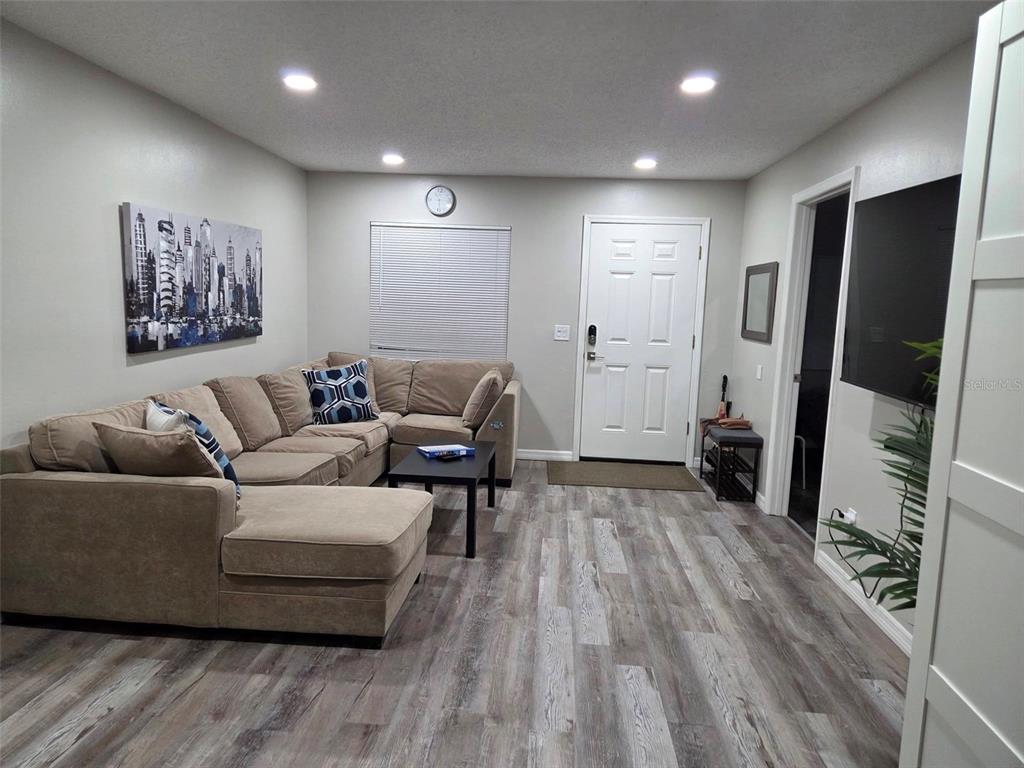 A living room features a tan sectional sofa, wood-look flooring, and a wall-mounted television — 8024 CANDLEWOODE DRIVE, LARGO, FL