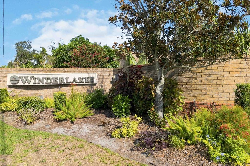 A brick wall features a sign for Winderlakes surrounded by various shrubs and a tree — 7813 SARANAC COURT, ORLANDO, FL