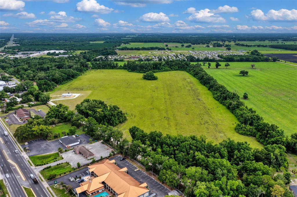 An aerial view shows a large, open grassy field bordered by trees and commercial buildings — Block L NW 11TH DRIVE, CHIEFLAND, FL