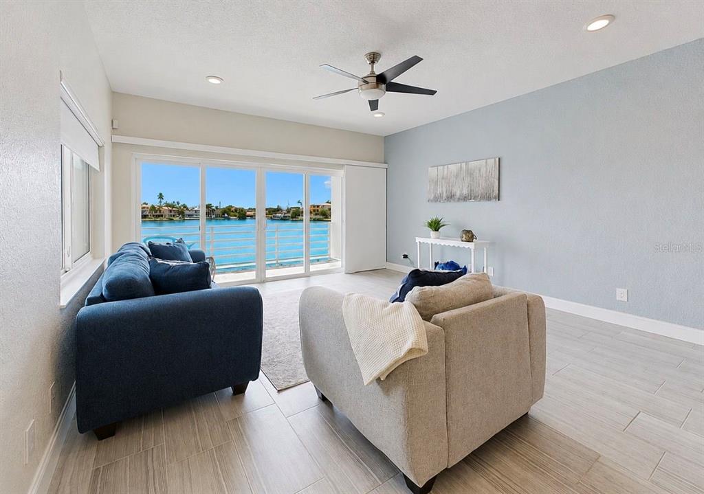 A living room features light tile flooring, a blue sofa, and sliding glass doors overlooking water — 4639 MIRABELLA COURT, ST PETE BEACH, FL