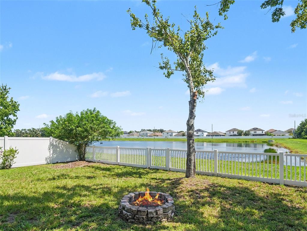 A backyard features a stone fire pit, a white fence, and a tree overlooking a pond — 10413 AVIAN FORREST DRIVE, RIVERVIEW, FL