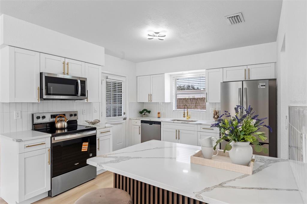 A modern kitchen features white cabinetry, stainless steel appliances, and a marble-topped island with seating — 230 BEVERLY BOULEVARD, BRANDON, FL
