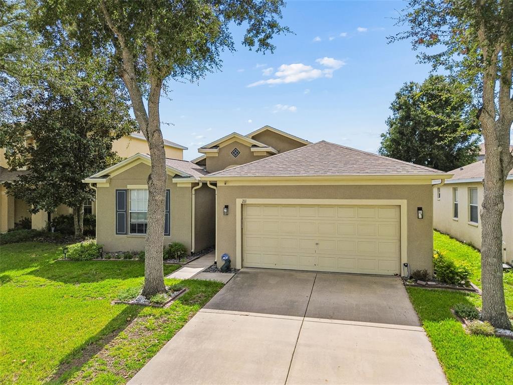A single-story suburban house with a tan stucco exterior, a two-car garage, and mature trees — 211 FAIRMONT DRIVE, SPRING HILL, FL