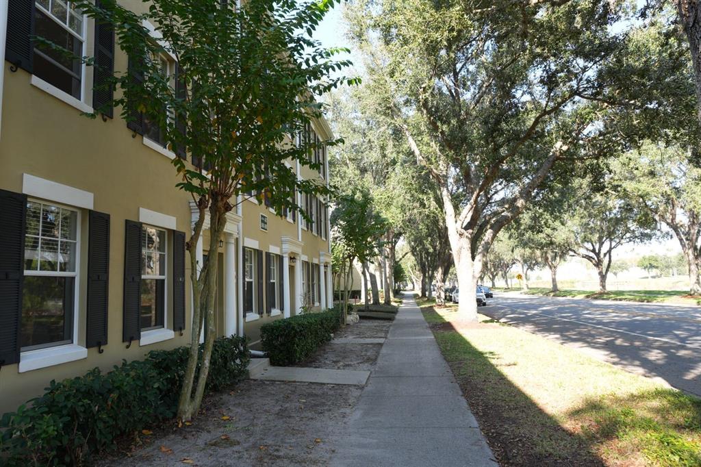 A sidewalk runs alongside a multi-story tan building with dark shutters and large oak trees — 1004 WATERSIDE DRIVE UNIT 1004, CELEBRATION, FL