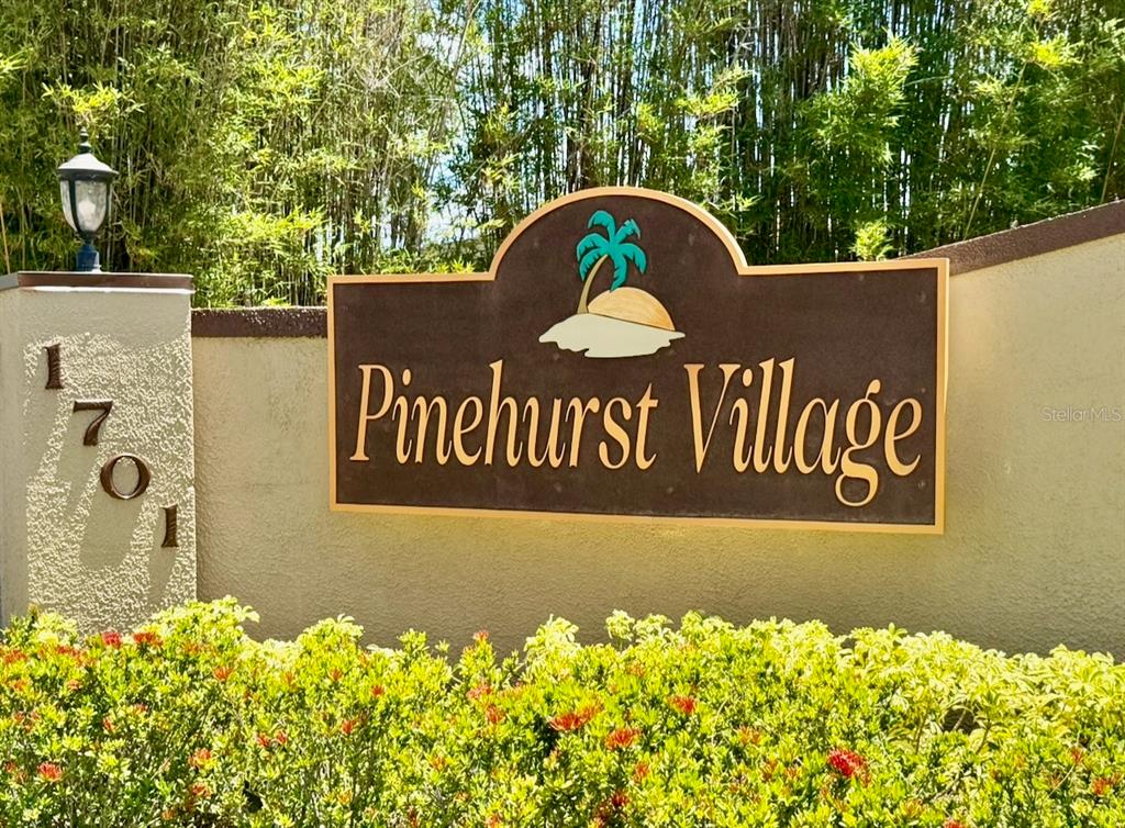 A sign for Pinehurst Village stands on a stucco wall next to a lamp post — 1701 PINEHURST ROAD UNIT 2F, DUNEDIN, FL