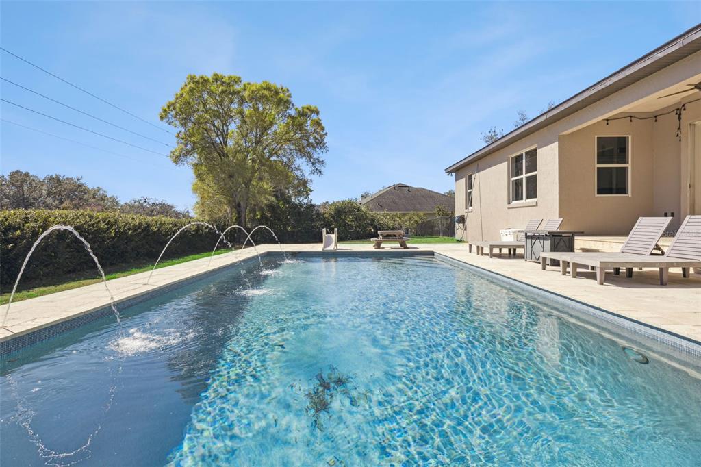 A rectangular swimming pool with water fountains sits on a patio beside a beige house — 11526 OSPREY POINTE BOULEVARD, CLERMONT, FL