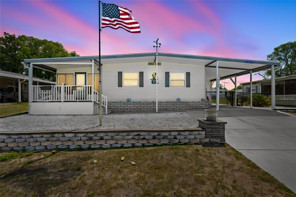 A single-story manufactured home with a front porch, carport, and American flag at sunset — 8979 HIGHPOINT BOULEVARD, BROOKSVILLE, FL