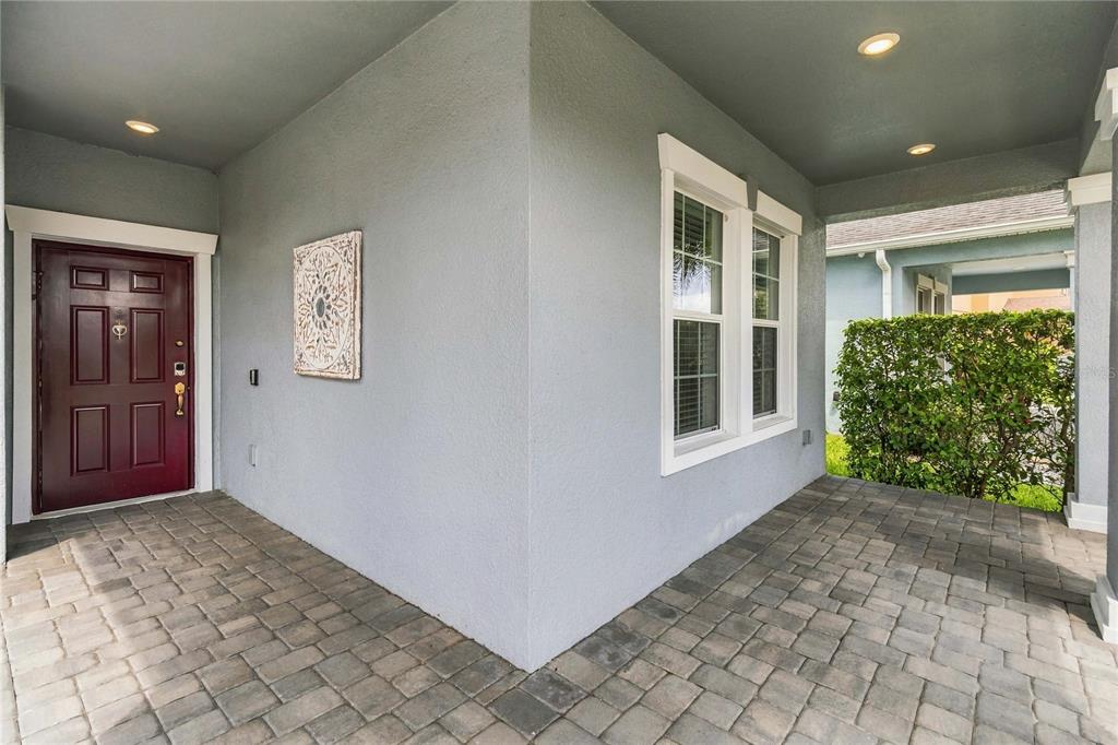 A covered front porch with gray stucco walls, a dark red door, and paver flooring — 3074 WHITE HORSE COURT, KISSIMMEE, FL