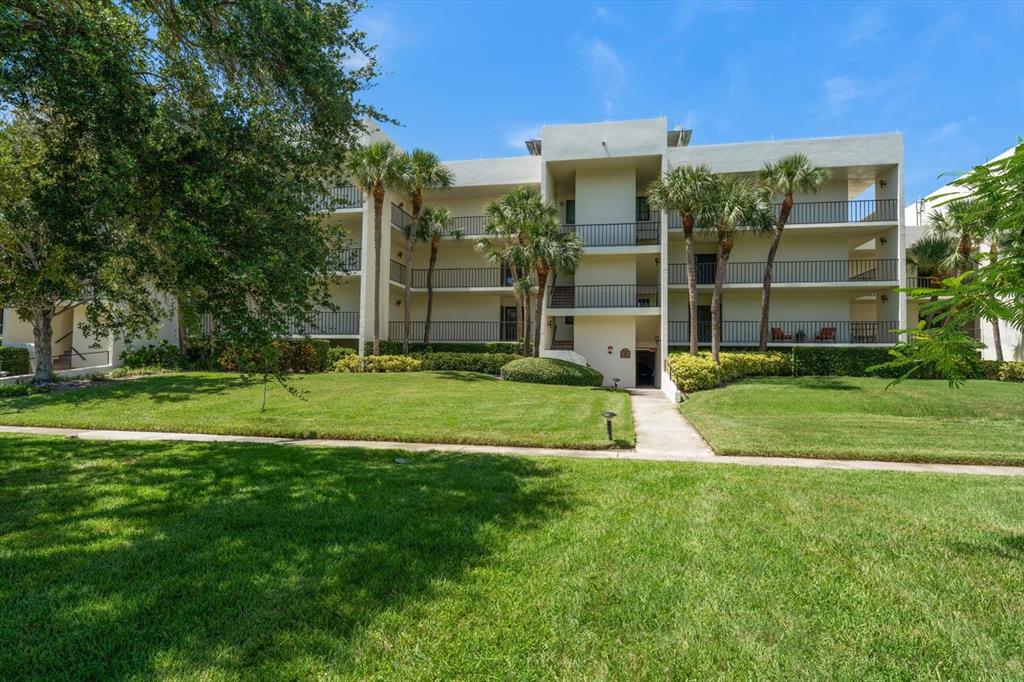 A three-story apartment building with balconies and palm trees sits behind a large grassy lawn — 50 COE ROAD UNIT 123, BELLEAIR, FL