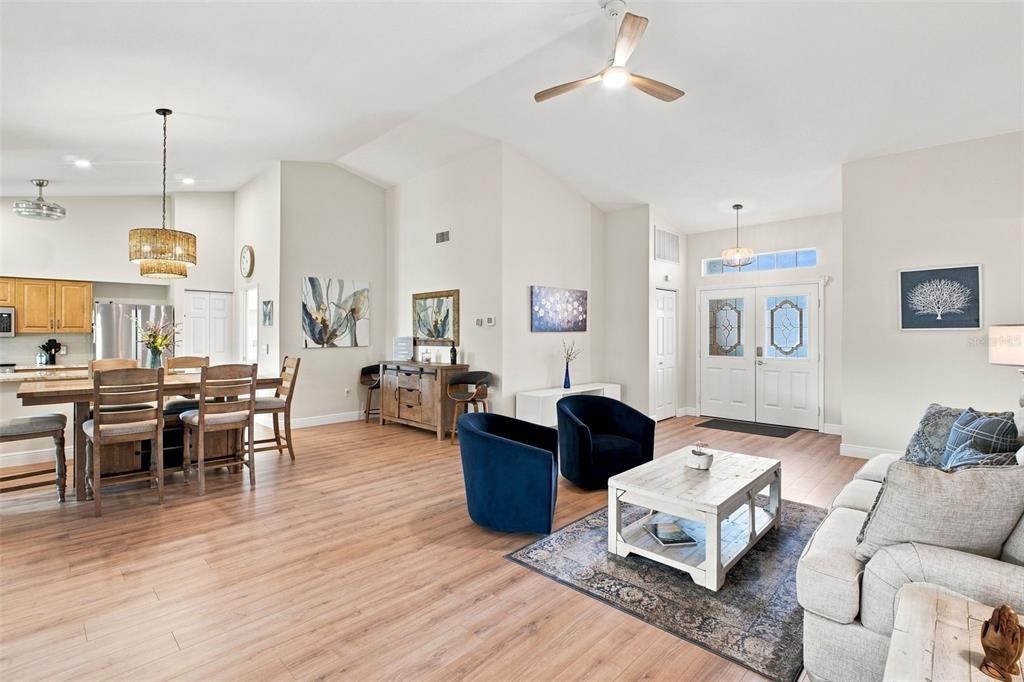 An open-concept living and dining area features light wood flooring, vaulted ceilings, and blue chairs — 1236 FLORA VISTA STREET, TRINITY, FL