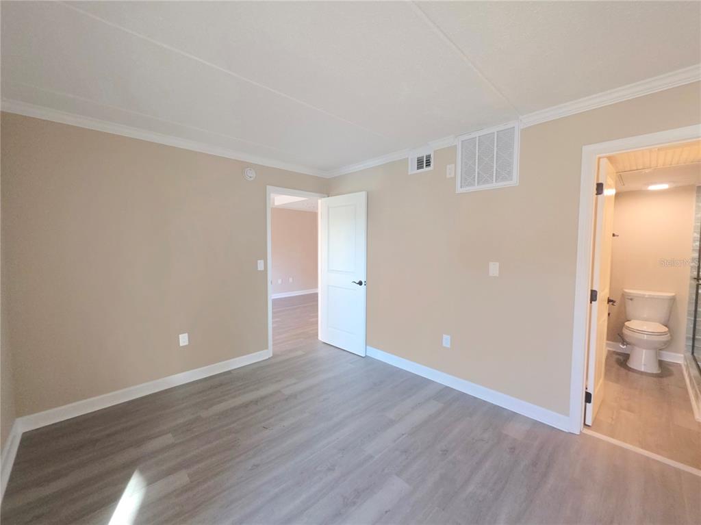 A bedroom with light-colored walls, gray wood-look flooring, an open doorway, and an attached bathroom — 8921 BLIND PASS ROAD UNIT 142, ST PETE BEACH, FL