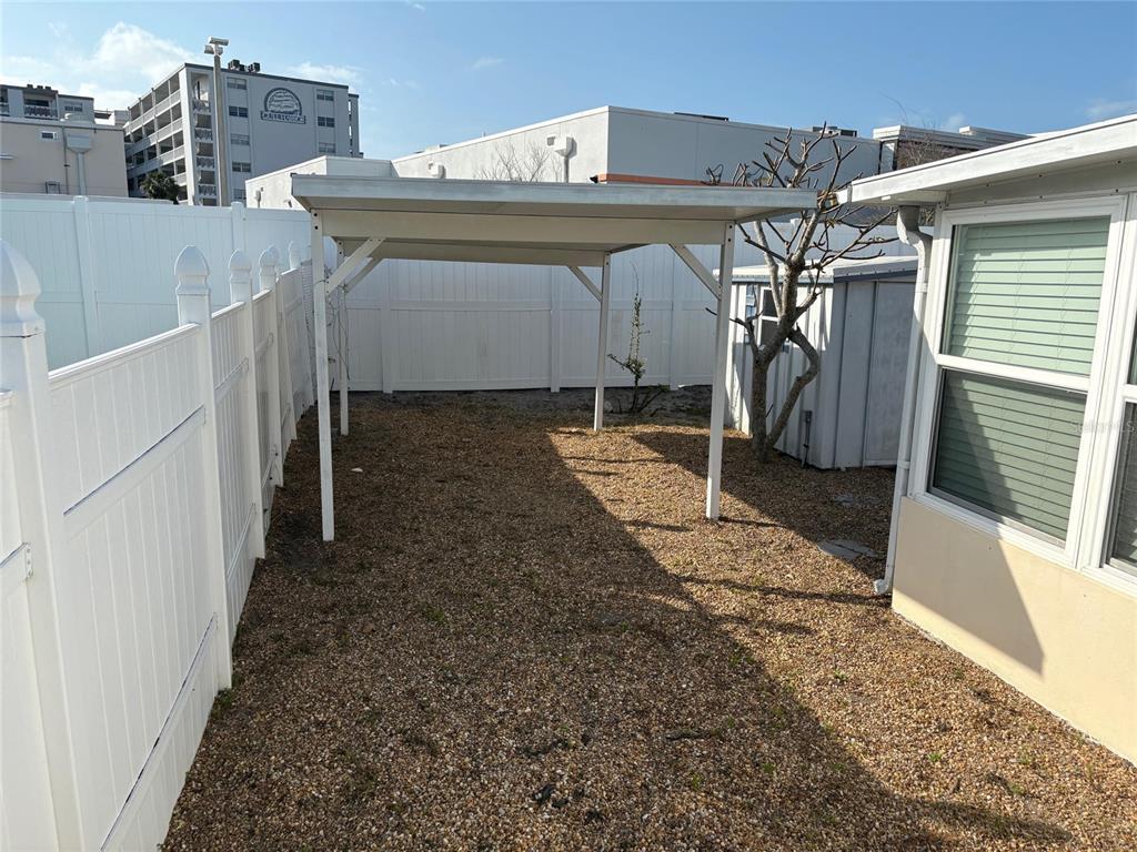 A gravel backyard features a white metal carport, a white vinyl fence, and a small tree — 111 174TH AVENUE E, REDINGTON SHORES, FL