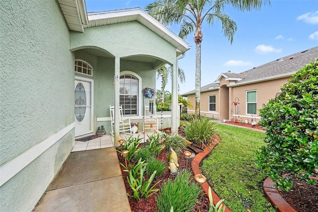 A light green house features a covered front porch with rocking chairs and landscaping — 12126 TOURNAMENT VIEW AVE, NEW PORT RICHEY, FL