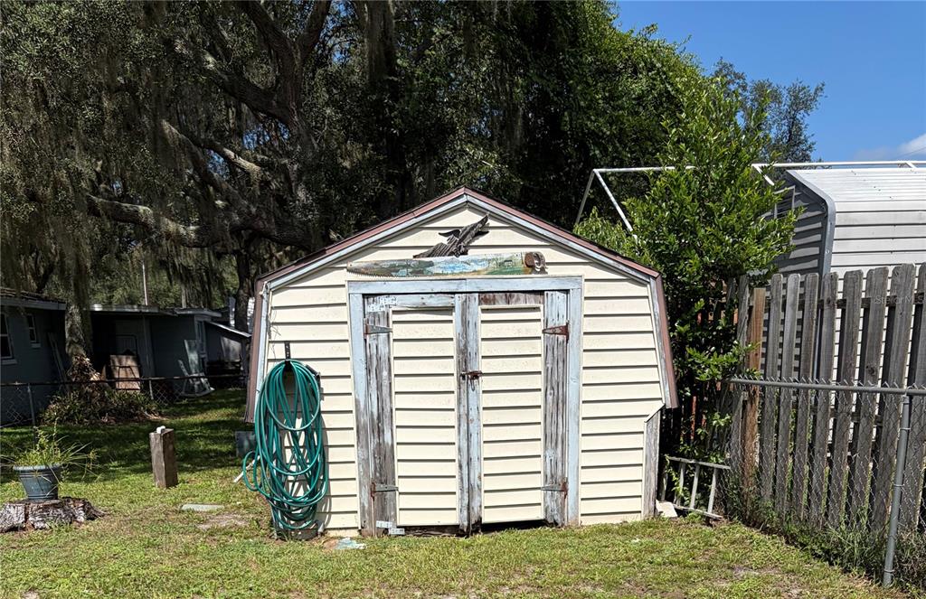 A light-colored storage shed with double doors stands in a grassy yard near a fence — 10138 E BASS CIRCLE, INVERNESS, FL