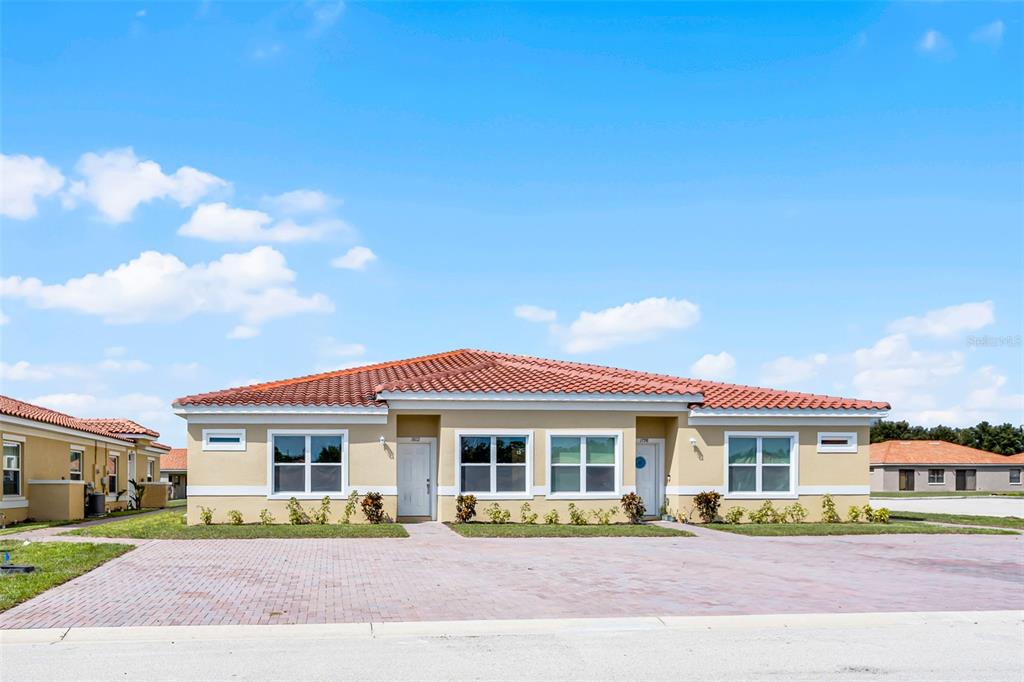 A single-story duplex with a tan stucco exterior, red tile roof, and brick paver driveway — 1826 CORIANDER DRIVE, KISSIMMEE, FL