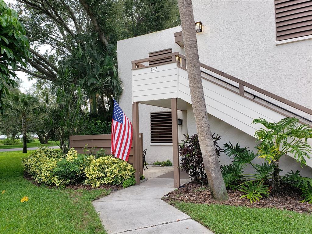 An exterior view of a residential building entrance with a staircase, American flag, and landscaping — 138 PINENEEDLE DRIVE, BRADENTON, FL