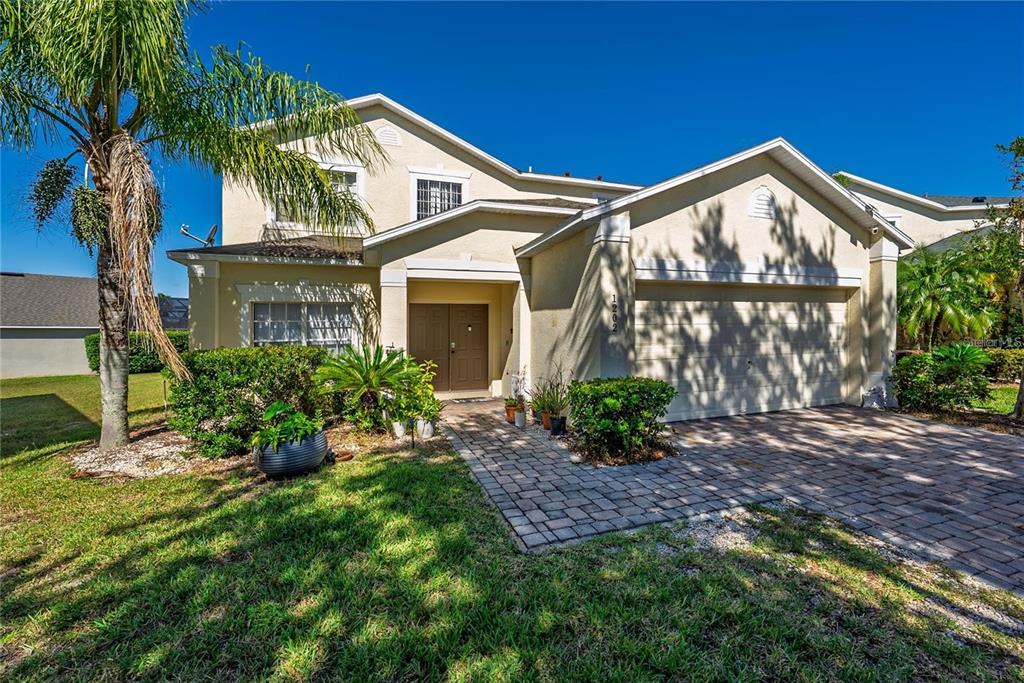 A two-story suburban house with a beige stucco exterior, a paved driveway, and a palm tree — 1202 CUMBRIAN LAKES COURT, KISSIMMEE, FL