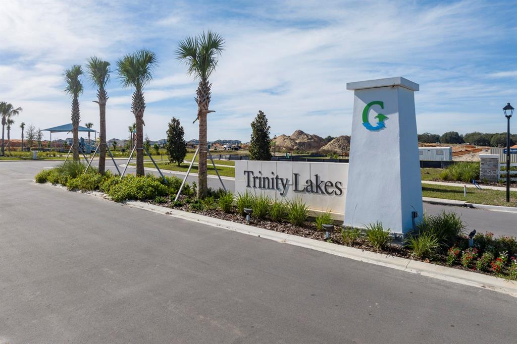 A white monument sign for Trinity Lakes stands beside a road with palm trees — 7040 ROLLING LEAF ROAD, GROVELAND, FL