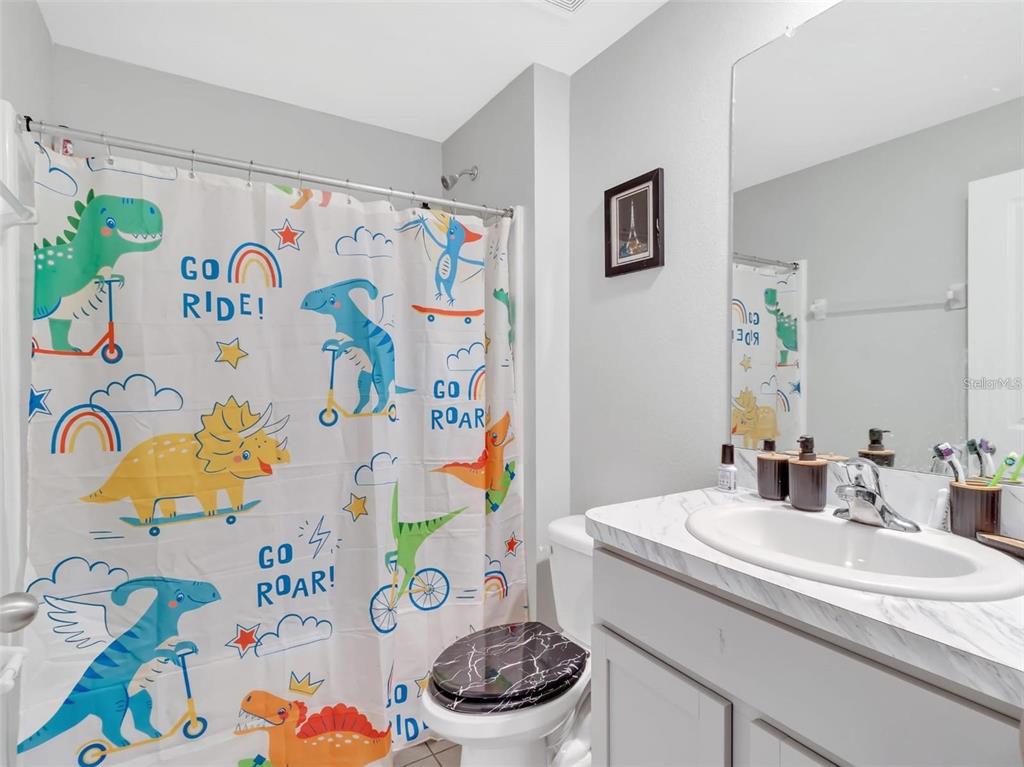 A bathroom features a dinosaur-themed shower curtain, a marble-patterned vanity, and a black toilet seat — 16636 WINDMILL FORGE PASS, WIMAUMA, FL