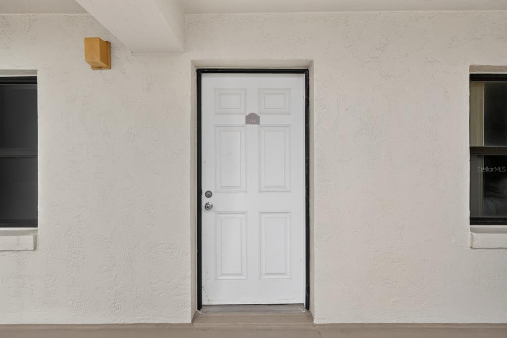 A white front door with a number plate is centered on a plain exterior wall — 2700 OCEAN SHORE BOULEVARD UNIT 114, ORMOND BEACH, FL