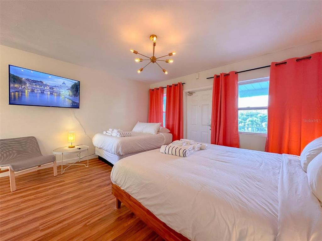 A bedroom features two beds, a wall-mounted television, a modern light fixture, and orange curtains — 333 HAMDEN DRIVE, CLEARWATER BEACH, FL