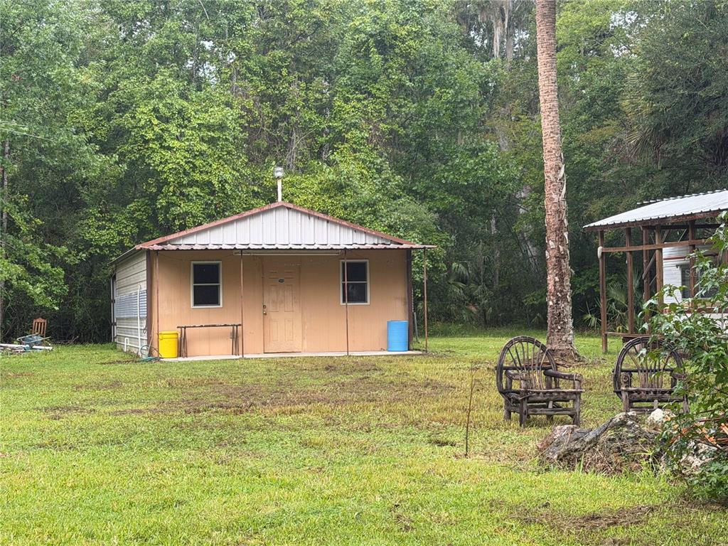 A small, tan-sided shed with a metal roof sits in a grassy, wooded yard — 17850 SE 32ND TERRACE, INGLIS, FL