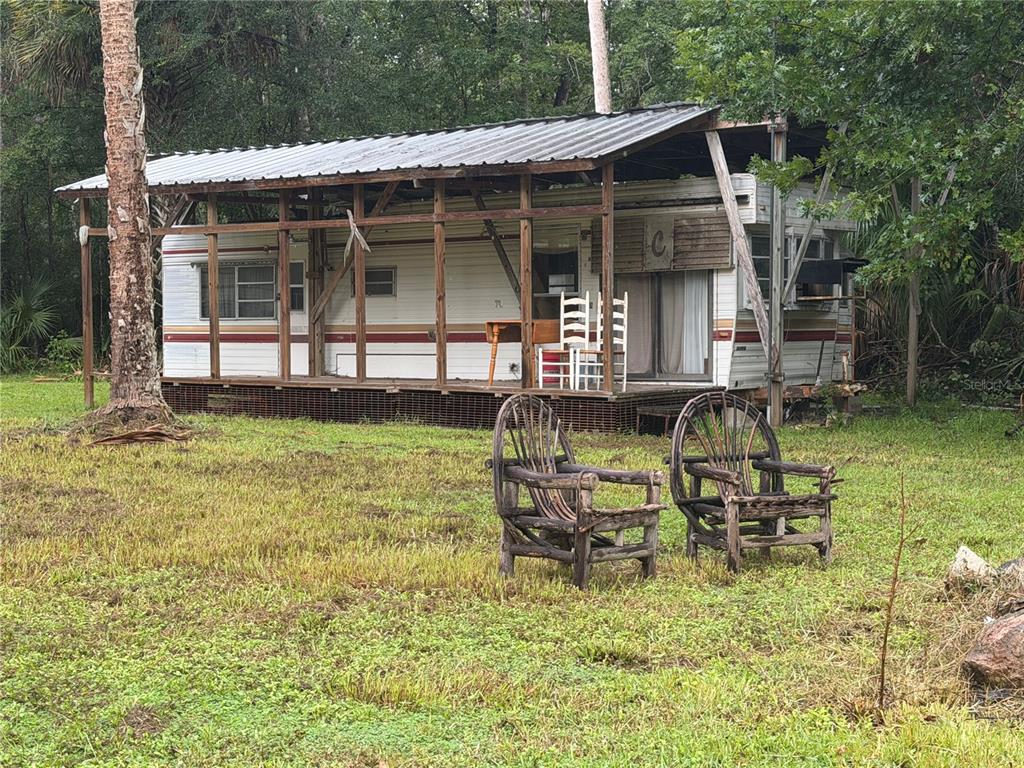 A weathered mobile home sits under a large metal roof structure in a grassy yard — 17850 SE 32ND TERRACE, INGLIS, FL
