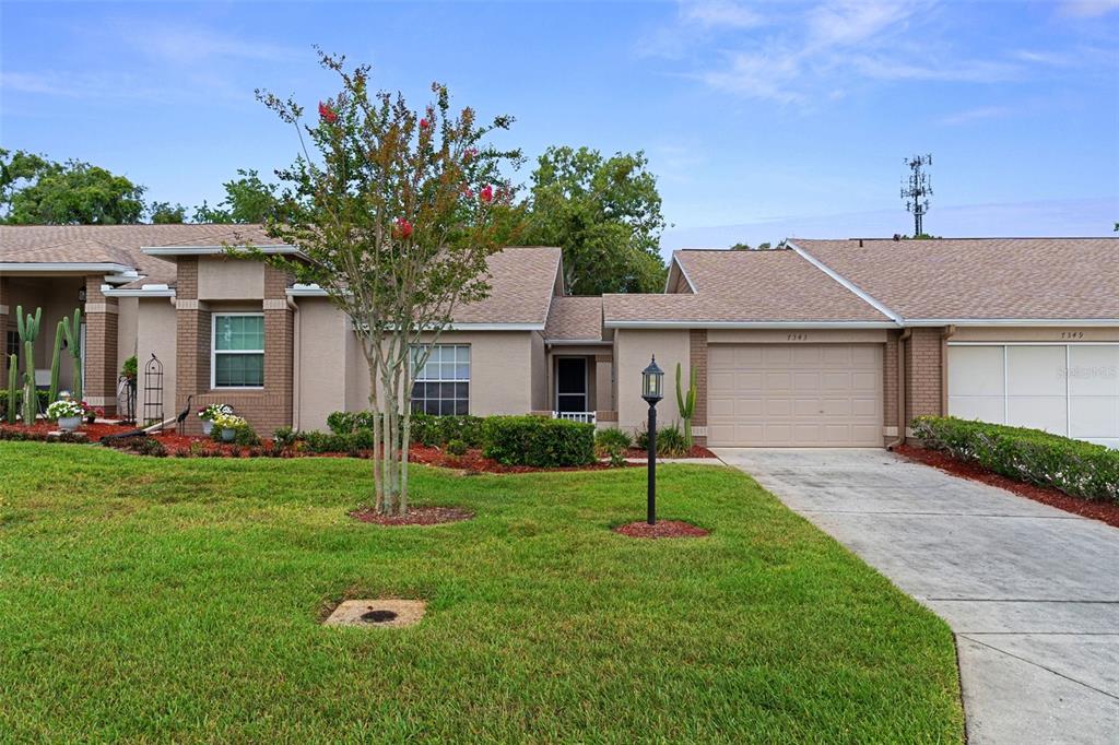 A single-story suburban house with a tan stucco exterior, a two-car garage, and a lawn — 7343 WILLOW BROOK DRIVE, SPRING HILL, FL