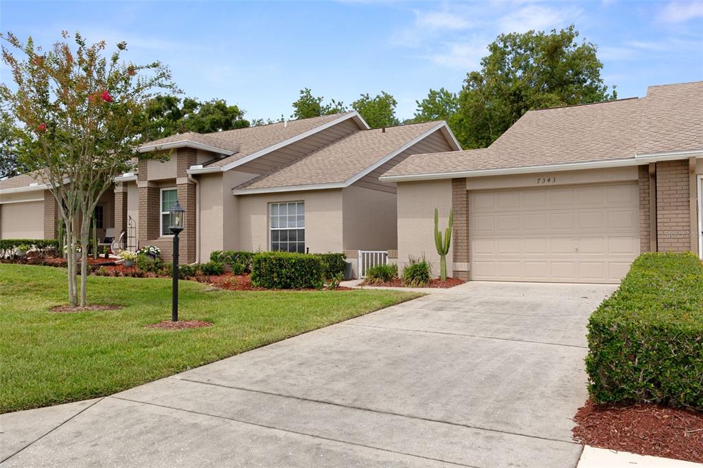 A tan single-story house with a two-car garage, a concrete driveway, and a manicured lawn — 7343 WILLOW BROOK DRIVE, SPRING HILL, FL