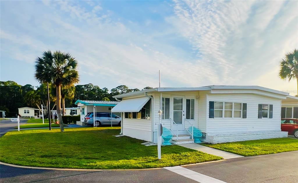 A white manufactured home with an attached carport sits on a grassy corner lot — 2282 PIAZZA DRIVE UNIT 117, SARASOTA, FL