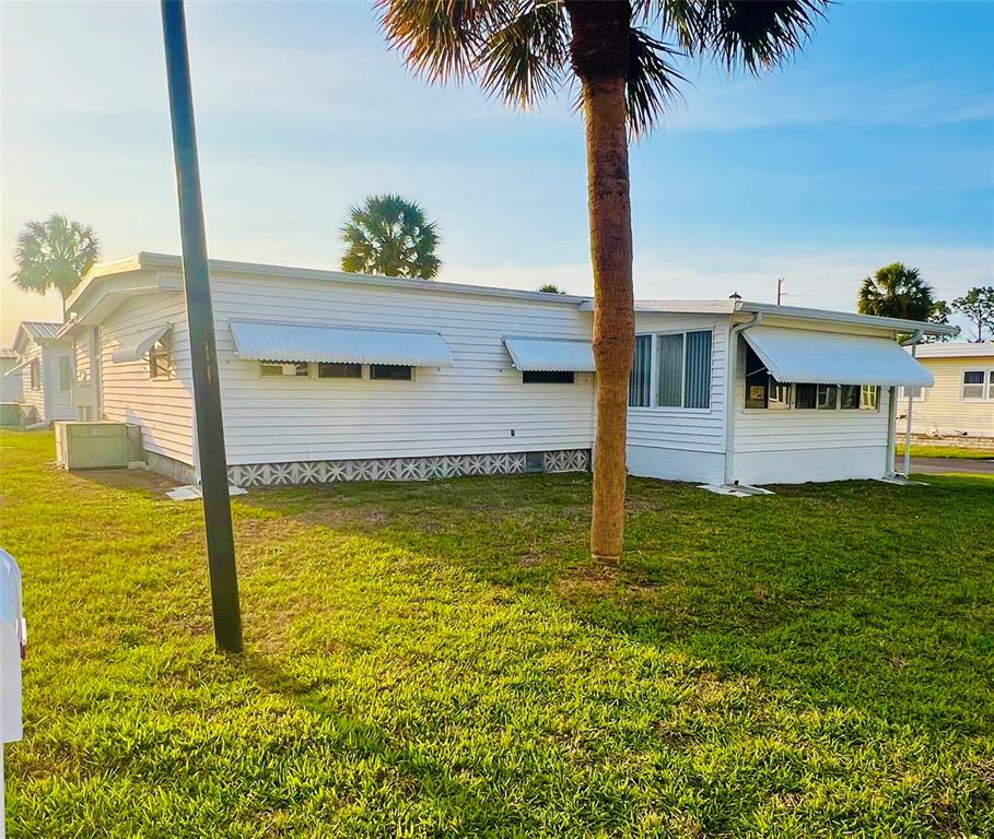 A white mobile home with metal awnings stands on a grassy lot with palm trees — 2282 PIAZZA DRIVE UNIT 117, SARASOTA, FL