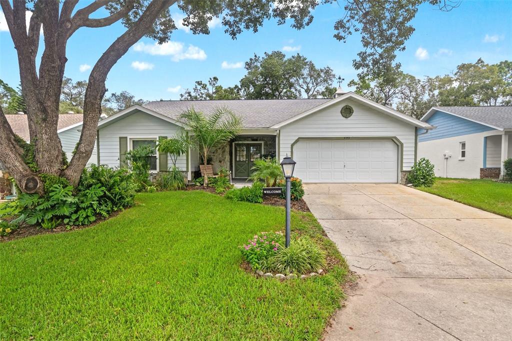 A single-story house with light siding, a two-car garage, and a large front yard tree — 6342 PINE MEADOWS DRIVE, SPRING HILL, FL