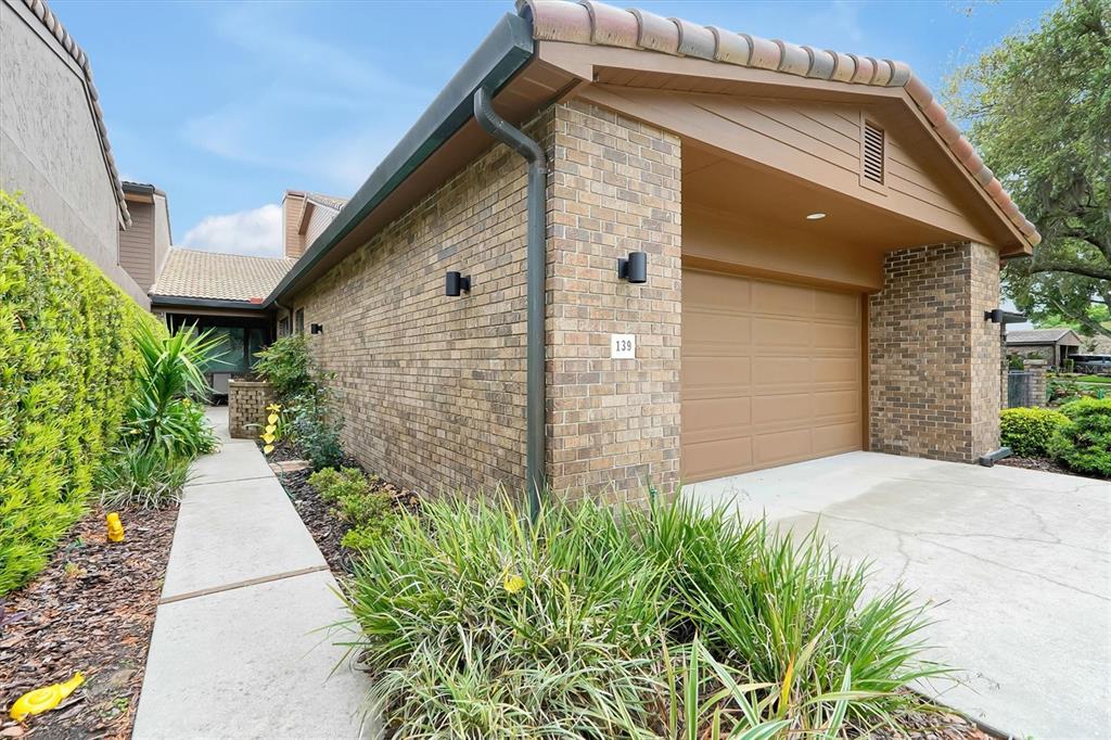 The exterior of a brick house features a garage, a concrete walkway, and landscaped greenery — 139 WODEN WAY, WINTER HAVEN, FL