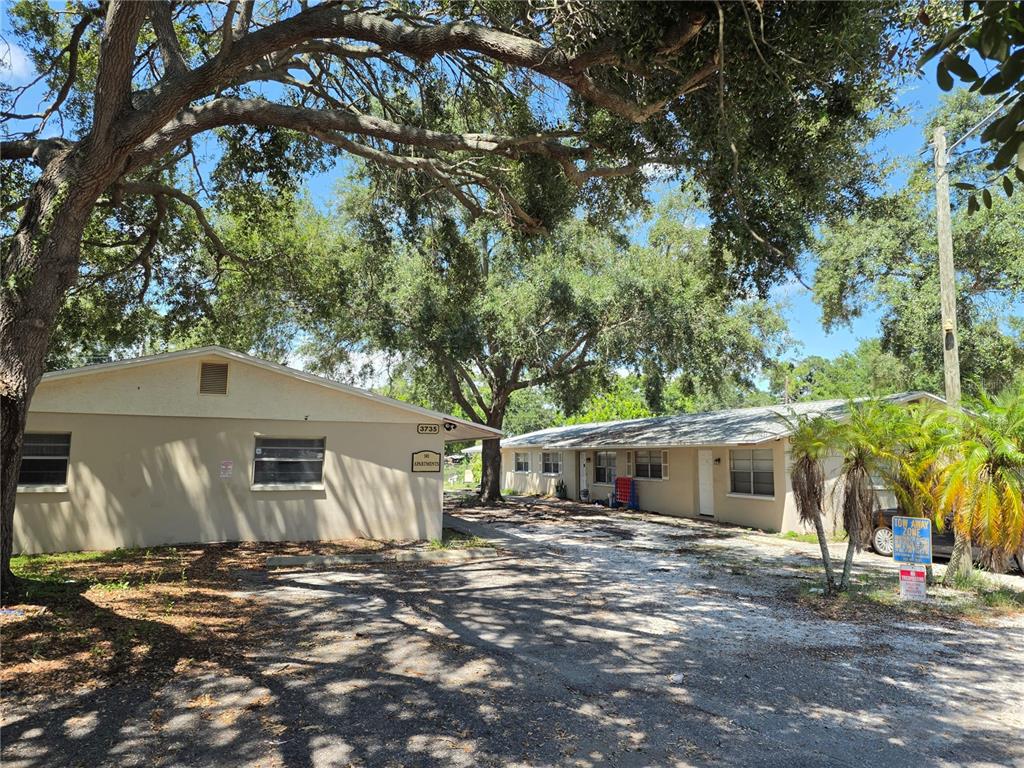 A gravel parking area sits in front of two single-story apartment buildings under large trees — 3735 141ST PLACE, LARGO, FL