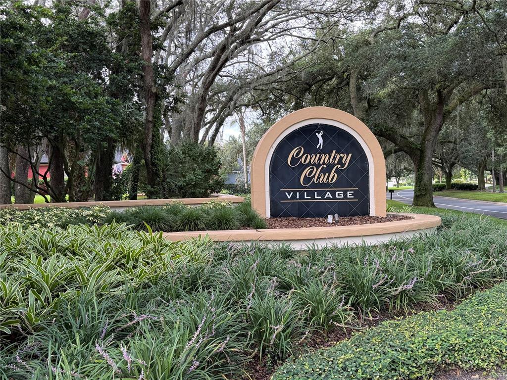 A curved stone sign for Country Club Village sits in a landscaped garden with trees — 1162 E WINGED FOOT CIRCLE, WINTER SPRINGS, FL