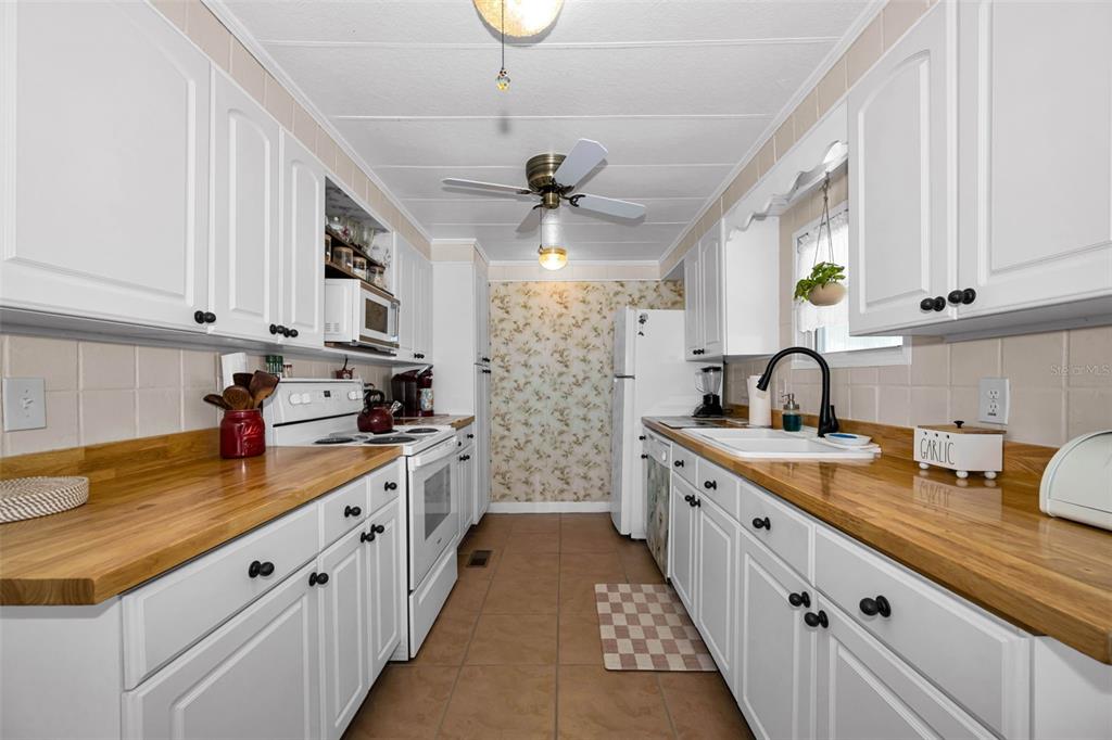 A galley kitchen features white cabinets, wood countertops, tile flooring, and floral wallpaper at the end — 143 MELODY LANE, TARPON SPRINGS, FL