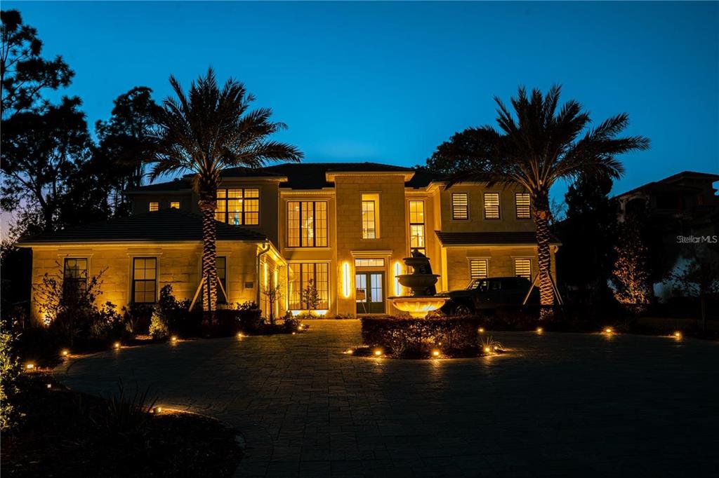 A two-story stone house at night features a paved driveway, fountain, and palm trees — 15926 PENDIO DRIVE, BELLA COLLINA, FL