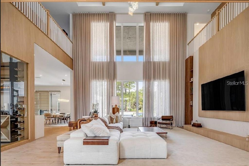 A double-height living room features light wood paneling, floor-to-ceiling curtains, and a large sectional sofa — 15926 PENDIO DRIVE, BELLA COLLINA, FL