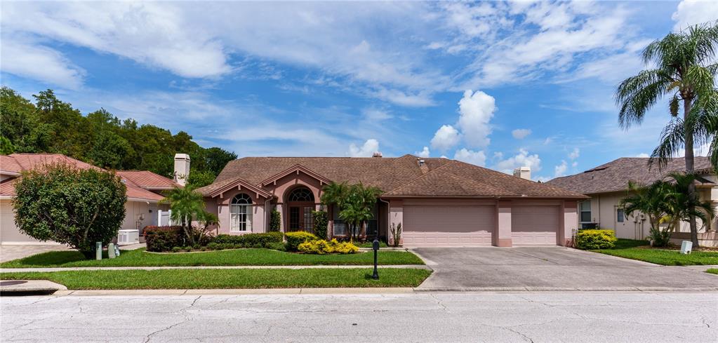 A single-story house with a brown shingled roof, beige stucco exterior, and a three-car garage — 7709 TIMBERWYCK COURT, NEW PORT RICHEY, FL
