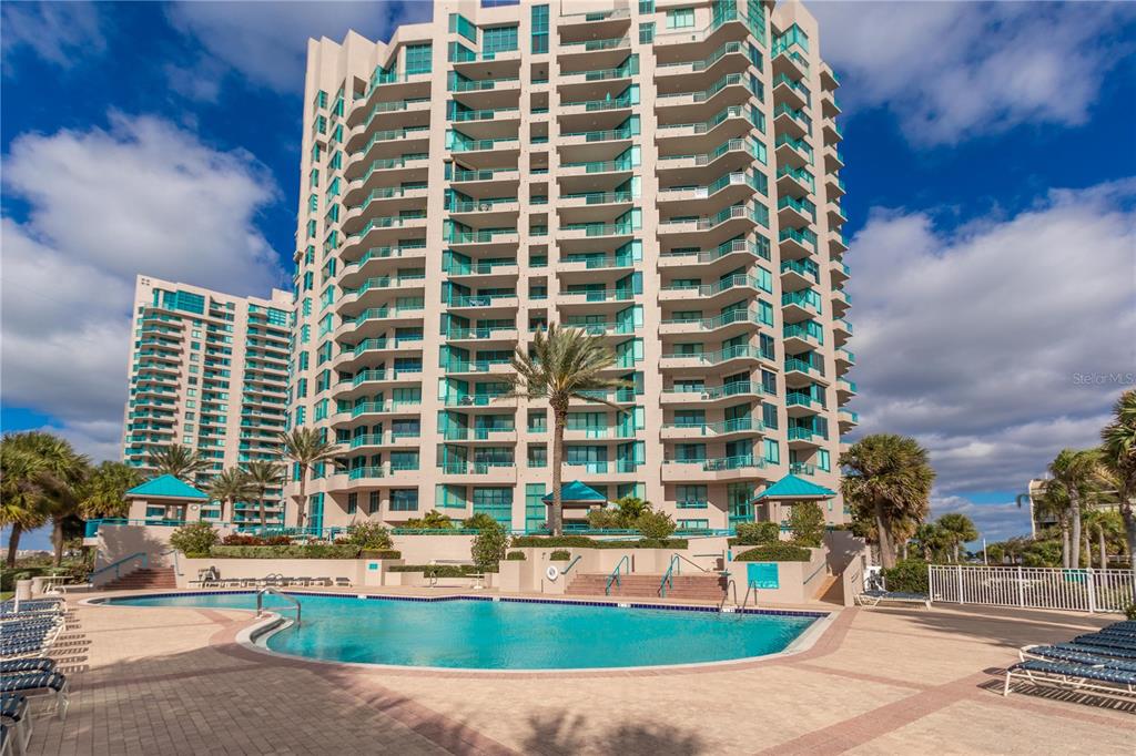 A large multi-story apartment building stands behind a swimming pool on a sunny day — 1560 GULF BOULEVARD UNIT 1607, CLEARWATER, FL