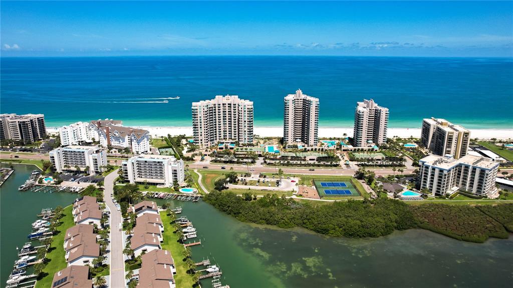 An aerial view shows beachfront high-rise condominiums, a marina, and tennis courts along the coast — 1560 GULF BOULEVARD UNIT 1607, CLEARWATER, FL
