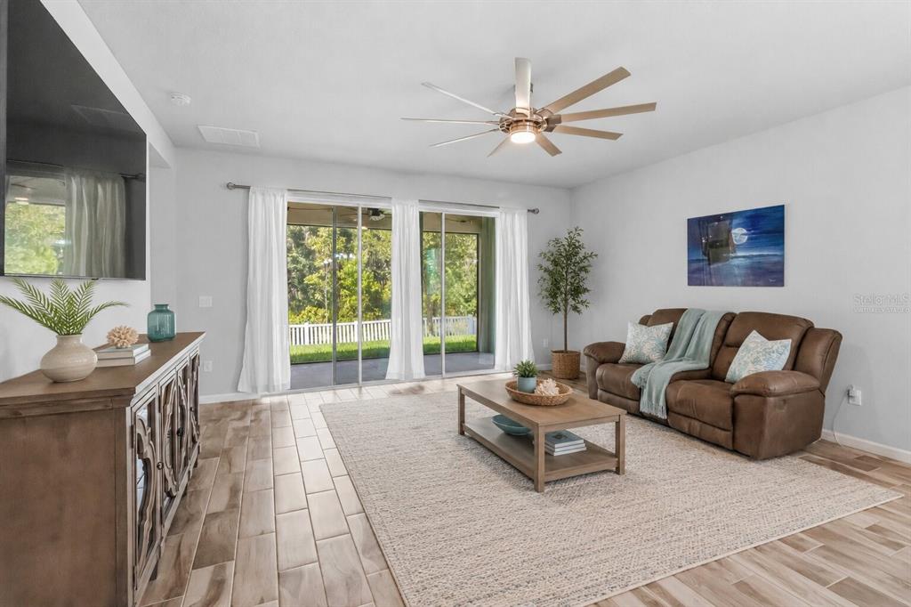 A living room with wood-look tile floors, a brown sofa, and sliding glass doors to a patio — 12943 SATIN LILY DRIVE, RIVERVIEW, FL