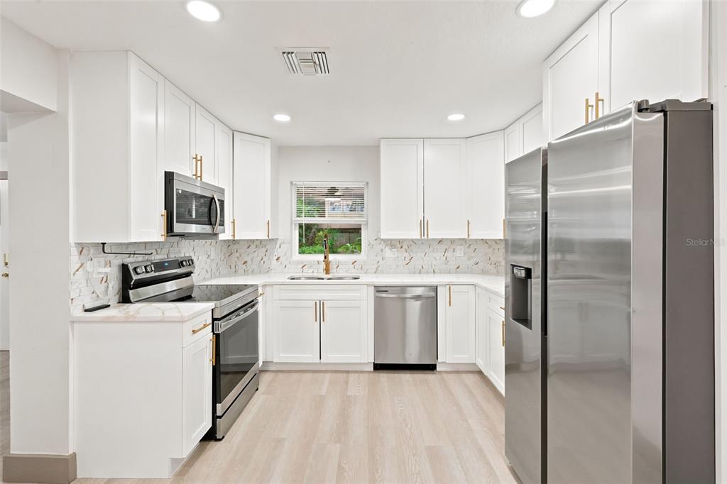An empty living room with light wood-look flooring, white walls, and a view into the kitchen — 6691 56TH AVENUE N, ST PETERSBURG, FL