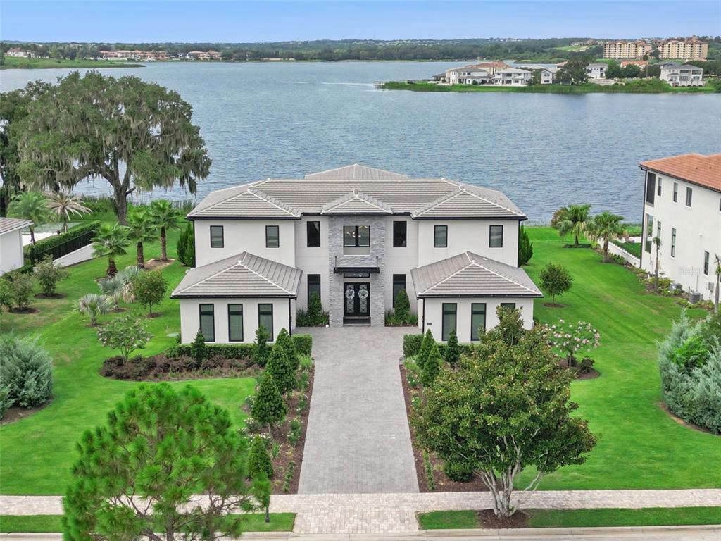 A large, two-story modern house with a stone facade sits on a lawn by a lake — 15421 PENDIO DRIVE, MONTVERDE, FL