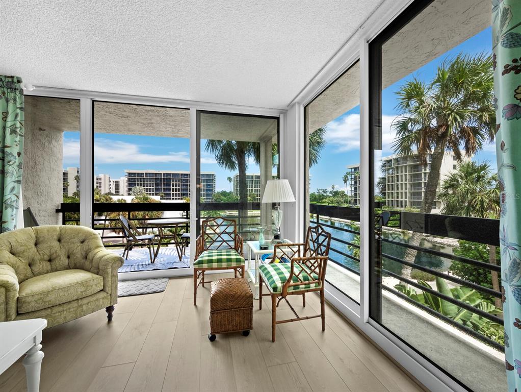 A sunroom features light wood flooring, a tufted armchair, and glass walls overlooking a canal — 1075 GULF OF MEXICO DRIVE UNIT 205, LONGBOAT KEY, FL