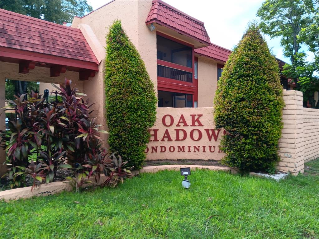 The exterior of a tan stucco condominium complex features a sign, landscaping, and red roof tiles — 2860 N POWERS DRIVE UNIT 8, ORLANDO, FL
