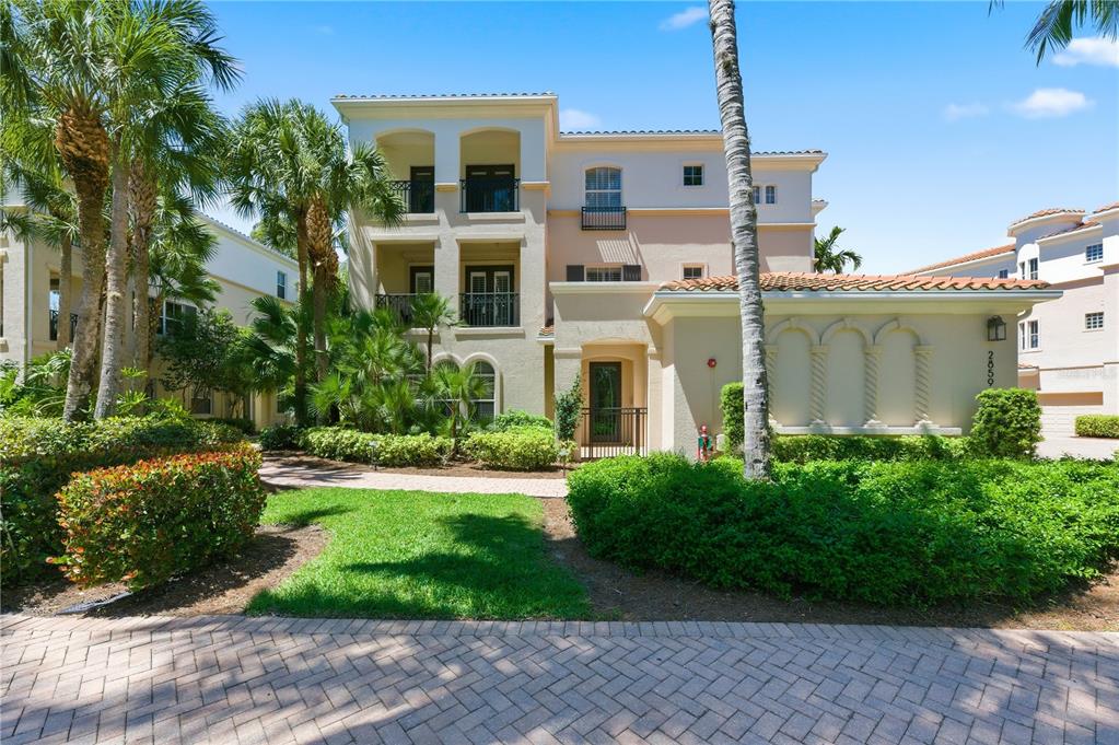 A three-story Mediterranean-style building features a paved walkway, lush landscaping, and palm trees — 2859 TIBURON BOULEVARD E UNIT 102, NAPLES, FL