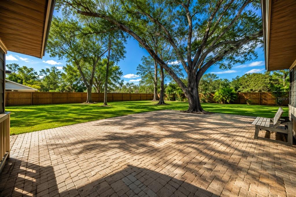 A paved patio overlooks a large, grassy backyard enclosed by a wooden fence with trees — 9502 59TH AVENUE E, BRADENTON, FL