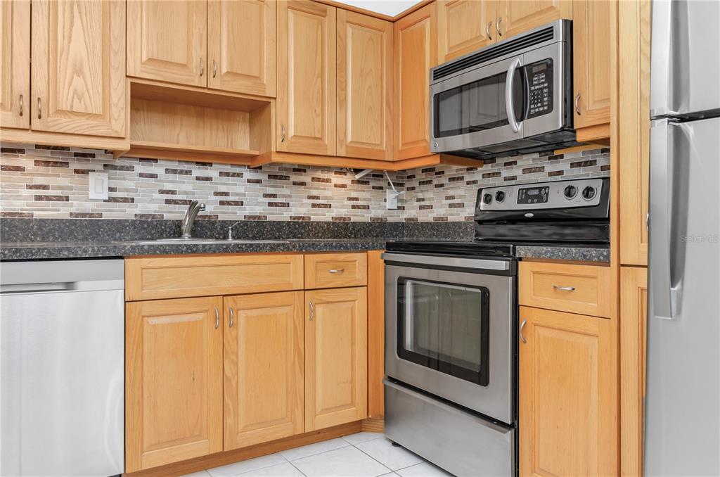 A kitchen features light wood cabinets, stainless steel appliances, and a mosaic tile backsplash — 3300 FOX CHASE CIRCLE N UNIT 231, PALM HARBOR, FL
