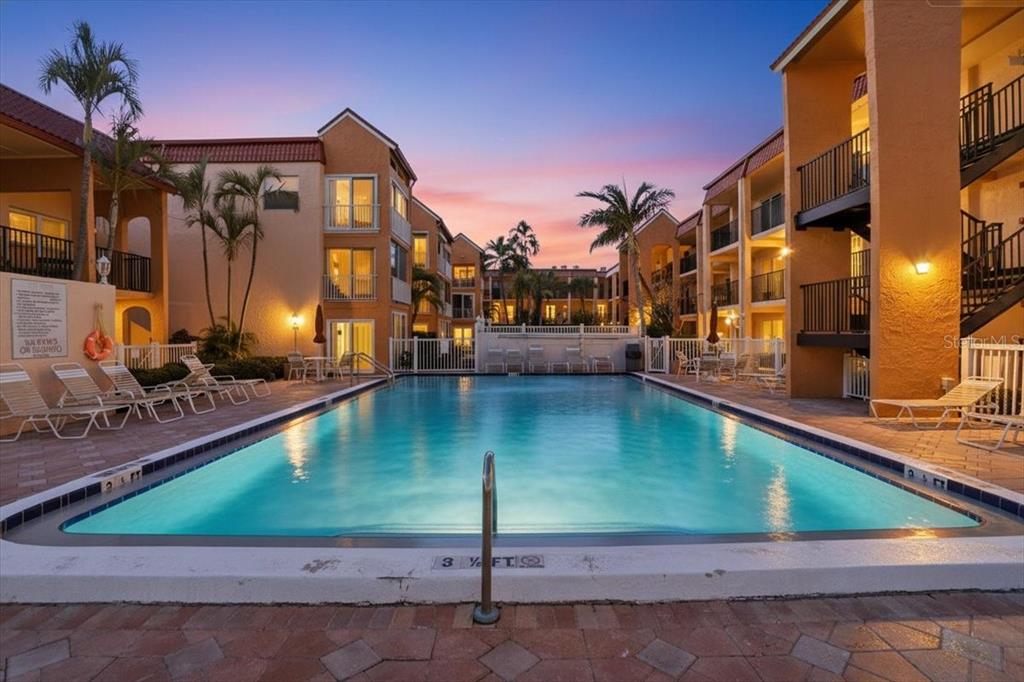 A rectangular swimming pool sits in a courtyard surrounded by multi-story apartment buildings at sunset — 8911 BLIND PASS ROAD UNIT 118, ST PETE BEACH, FL